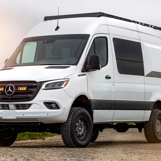 A white Mercedes-Benz Sprinter van with black fenders and off-road tires, parked outdoors.