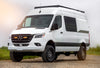 A white Mercedes-Benz Sprinter van with black fenders and off-road tires, parked outdoors.