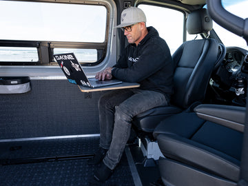 A person seated in a Sprinter van with a laptop on their lap, showcasing the interior and potential use of the VANNON Lagun B-Pillar Mount.