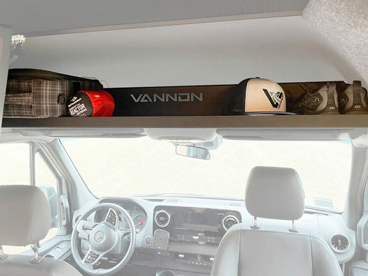 Overhead shelf installed inside the cabin of a van, made of aluminum with a padded EVA foam liner, holding various items such as a red and white checkered bag and a white helmet.