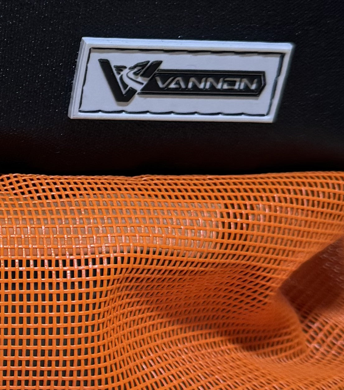 Close-up of a VANNON logo on a black background with orange mesh fabric.